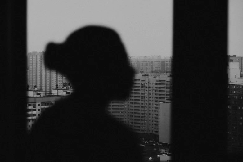 a black and white photo of a person looking out a window, a black and white photo, by Shang Xi, city background in silhouette, xiaoguang sun, close - up portrait shot, the photo was taken from afar