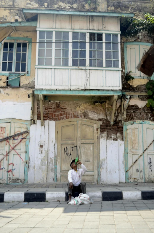 a man sitting on the sidewalk in front of a building, inspired by Altoon Sultan, flickr, derelict house, indonesia, colonial style, white building