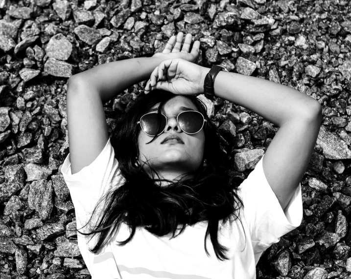 a woman laying on top of a pile of rocks, a black and white photo, pexels, realism, with sunglass, indian girl with brown skin, dressed in a white t shirt, trending photo