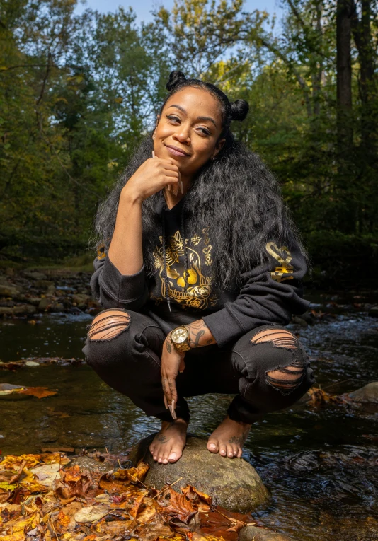 a woman sitting on top of a rock next to a river, rapper, alexis franklin, promotional image, golden glow