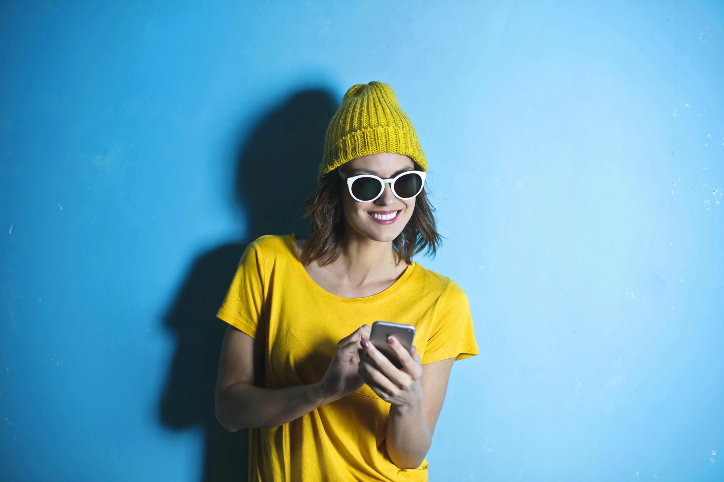 a woman standing in front of a blue wall holding a cell phone, wearing sunglasses and a cap, yellow hue, hipster, : :