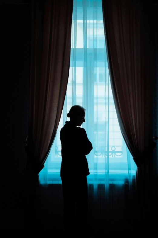 a silhouette of a person standing in front of a window, inspired by Nan Goldin, unsplash, romanticism, blue room, curtains, crossed arms, looking sad