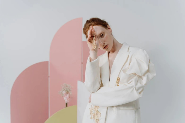 a woman in a white robe standing in front of a pink wall, inspired by Russell Dongjun Lu, trending on pexels, aestheticism, woman with red hair, gold suit, wearing futuristic white suit, ivory rococo