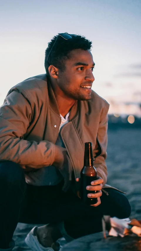 a man sitting next to a fire with a bottle of beer, by Cosmo Alexander, pexels contest winner, wearing a bomber jacket, tanned skintone, smiling male, drew hill