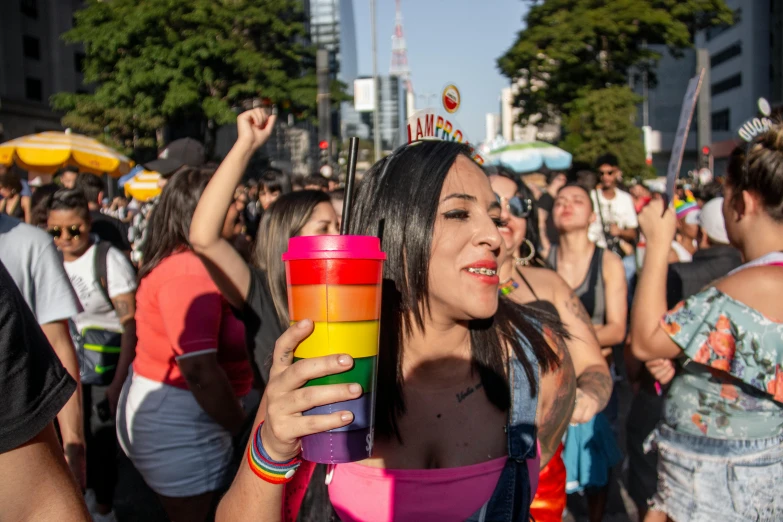 a woman holding a cup in front of a crowd of people, rainbow tubing, avenida paulista, other women dancing behind, profile image