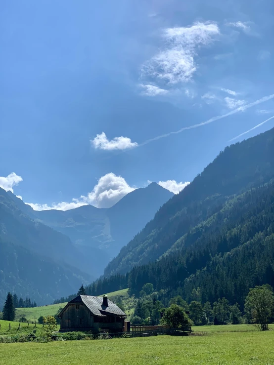 a house in a field with mountains in the background, by Anna Haifisch, pexels contest winner, les nabis, lush forest in valley below, profile image, clear blue skies, looking threatening