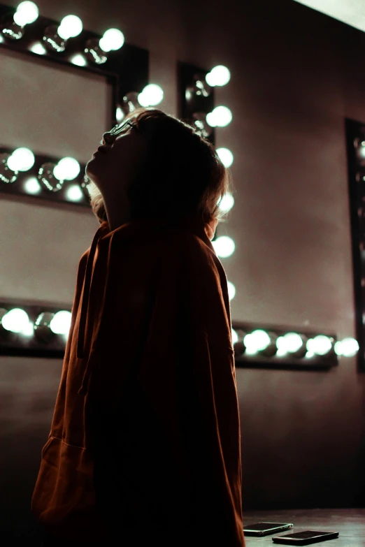 a woman standing in front of a lighted mirror, inspired by Elsa Bleda, pexels contest winner, red robe, ( ( stage lights ) ), ( ( theatrical ) ), facing away from camera