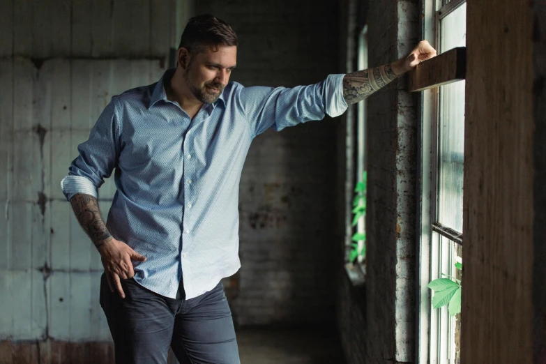 a man in a blue shirt leaning against a window, inspired by Paul Davis, pexels contest winner, symbolism, photograph of a sleeve tattoo, tailored clothing, rugged man portrait, walking to the right