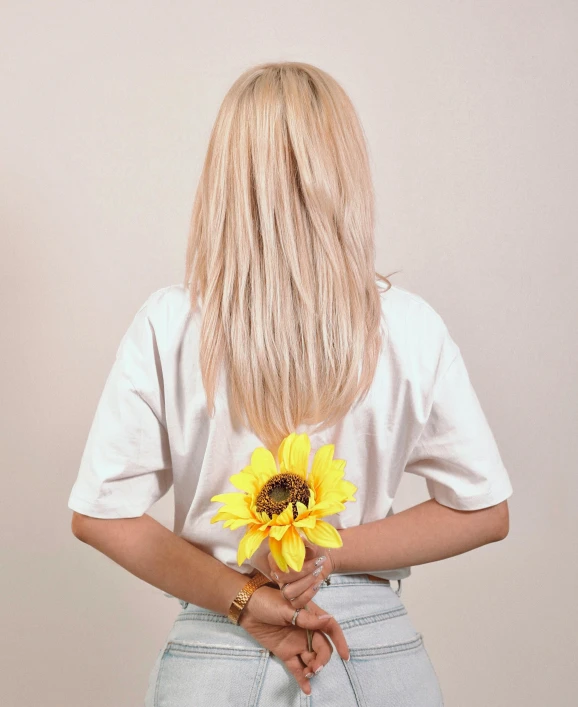 a woman holding a sunflower in her hands, an album cover, inspired by Elsa Bleda, dressed in a white t-shirt, back view », extra-light natural blonde hair, detailed product image