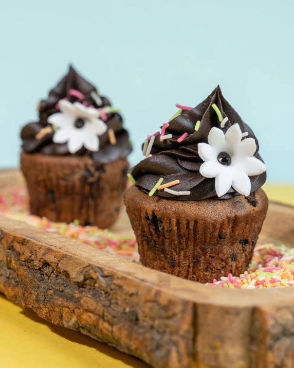 three cupcakes with chocolate frosting and sprinkles on a wooden tray, by Helen Stevenson, unsplash, flower power, sculpted, profile image, woodland location