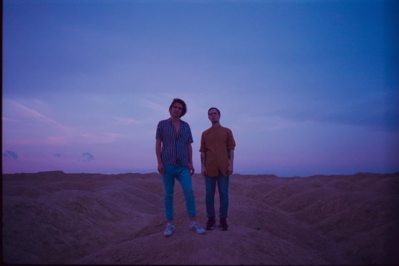 a couple of men standing on top of a sandy beach, an album cover, unsplash, antipodeans, desert colors, bisexual lighting, temples, looking towards camera