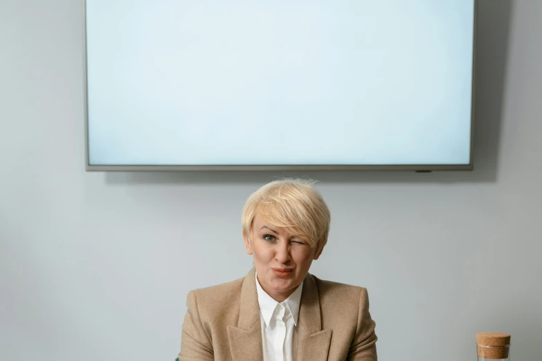 a woman sitting at a table in front of a tv, inspired by Cerith Wyn Evans, short blond hair, corporate portrait, in front of white back drop, androgynous person