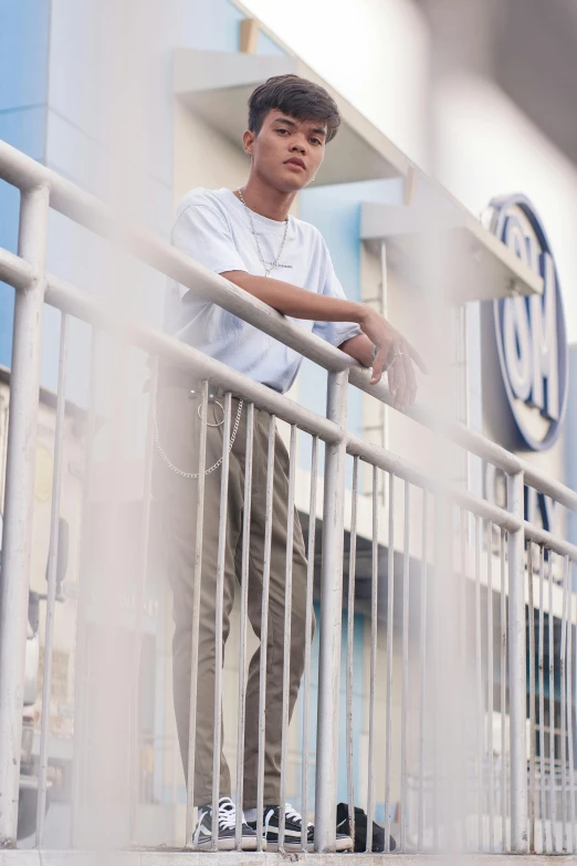 a man leaning on a railing in front of a building, she is wearing streetwear, white and pale blue, thawan duchanee, full frame image