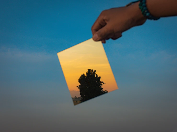 a person holding a piece of paper with a tree in the background, by Jan Rustem, pexels contest winner, abstract illusionism, refracted sunset, transparent glass surfaces, sky gradient, on a reflective gold plate