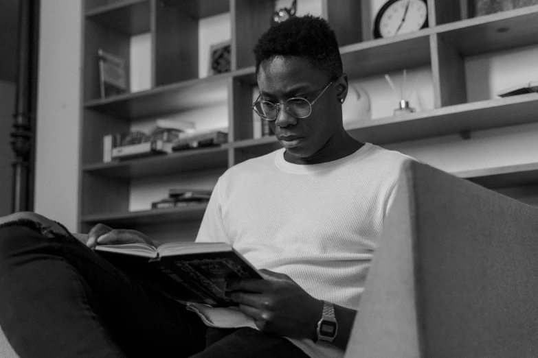 a man sitting on a couch reading a book, a black and white photo, pexels, adebanji alade, student, wearing thin large round glasses, sitting on a store shelf