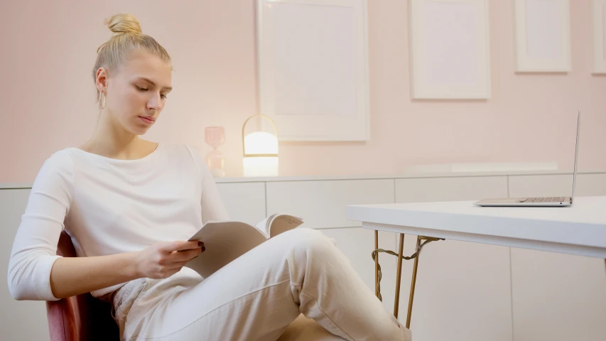 a woman sitting in a chair reading a book, by Liza Donnelly, trending on pexels, aestheticism, soft light 4 k in pink, on a white table, relaxed. gold background, kirsi salonen