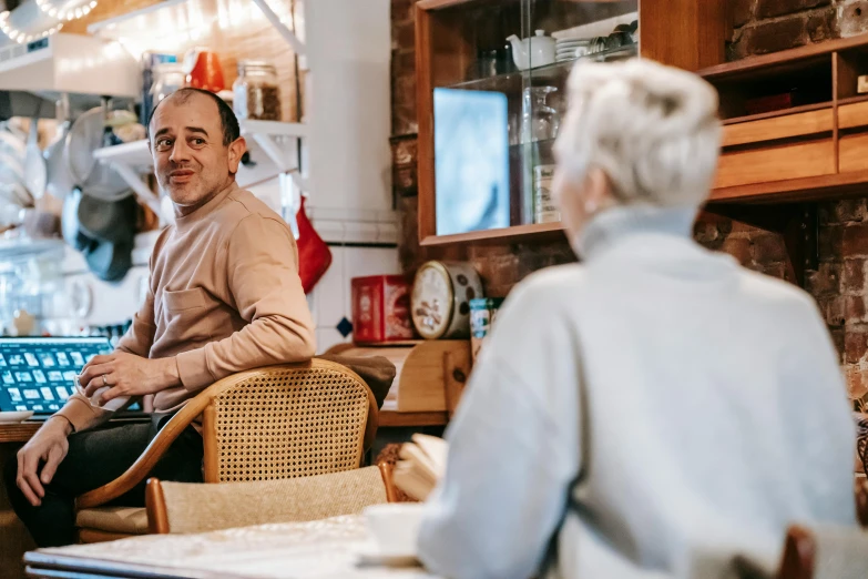 a man sitting in a chair next to a woman, by Lee Loughridge, pexels contest winner, happening, cozy cafe background, older male, welcoming grin, lachlan bailey