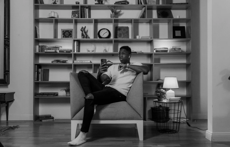 a black and white photo of a man sitting on a couch, by Marshall Arisman, pexels, hold up smartphone, black man, sitting on a store shelf, sitting in fancy chair