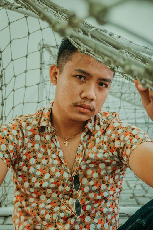 a man sitting in front of a fishing net, an album cover, inspired by Byron Galvez, unsplash, patterned clothing, lovingly looking at camera, impeccable military composure, lgbtq