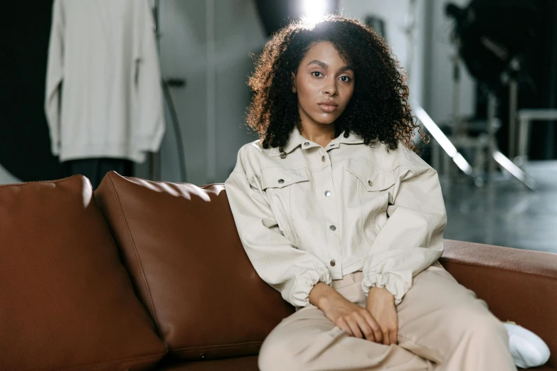 a woman sitting on top of a brown couch, inspired by Afewerk Tekle, trending on pexels, wearing off - white style, she wears a jacket, curly haired, utility