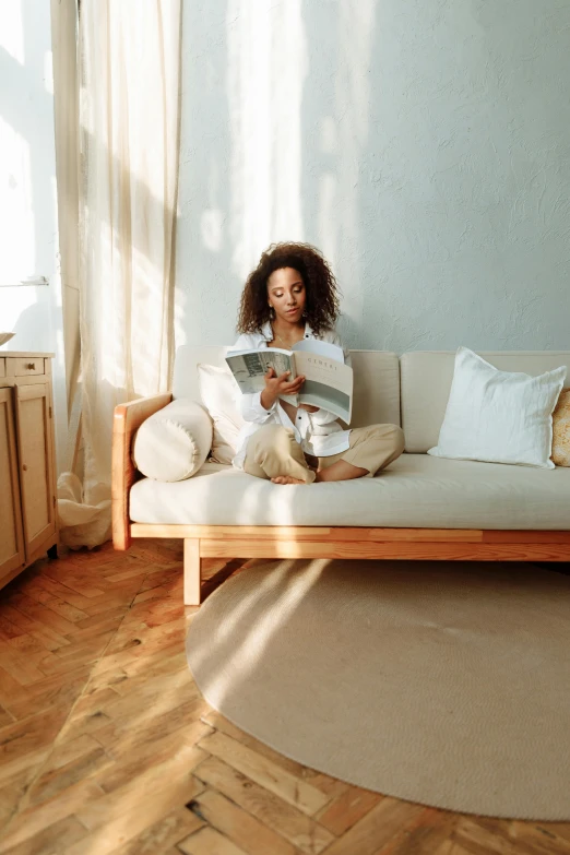 a woman sitting on a couch reading a book, diffuse natural sun lights, cream - colored room, millennial vibes, african american woman