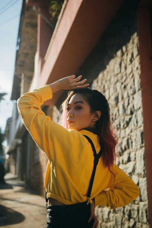 a woman standing in front of a brick wall, inspired by Elsa Bleda, trending on pexels, realism, wearing a yellow hoodie, vietnamese woman, hands in her hair, androgynous person