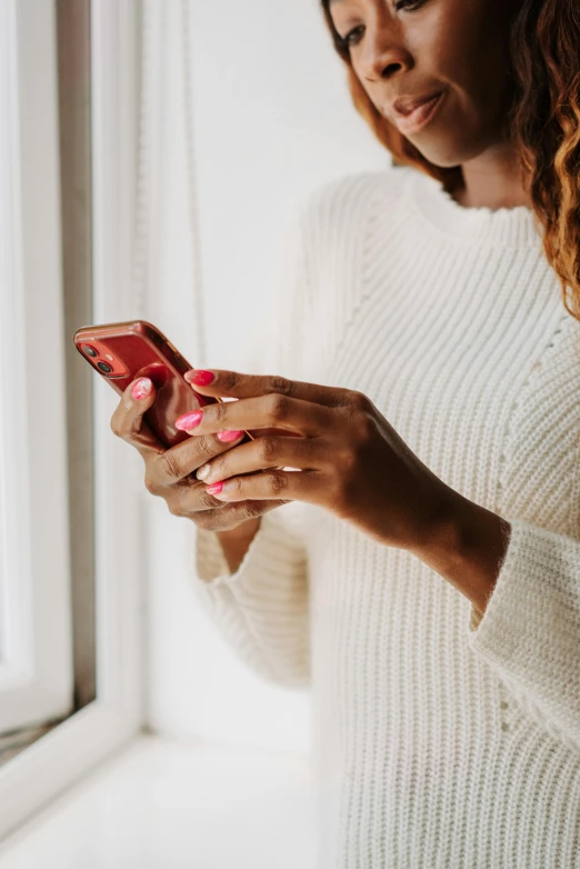 a woman standing in front of a window looking at her cell phone, trending on pexels, wearing a white sweater, dating app icon, pink hue, sleek hands