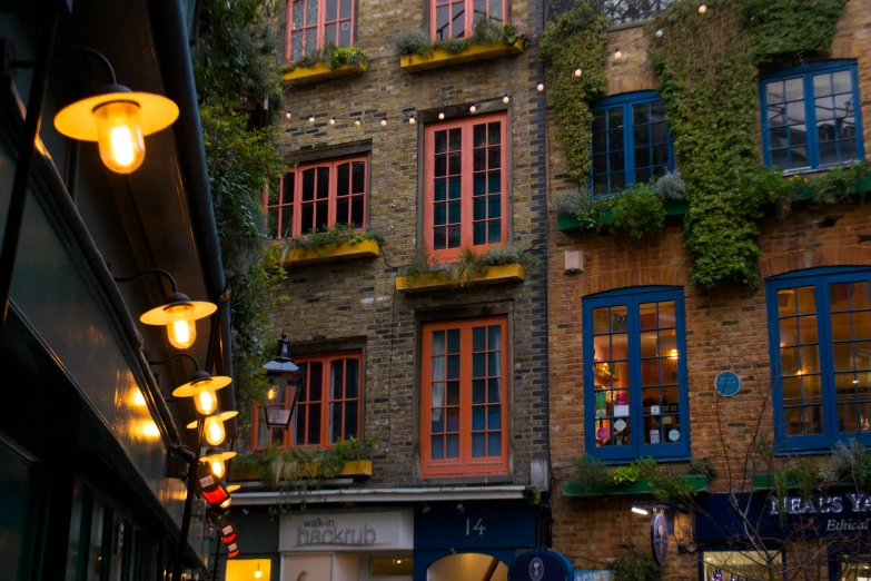 a group of people walking down a street next to tall buildings, vines on the walls, london at night, colorful caparisons, golden windows