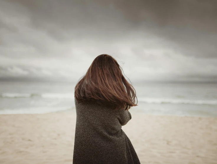 a woman standing on top of a sandy beach, by Lucia Peka, unsplash, realism, in a cloak with long hairs, overcast gray skies, girl with dark brown hair, covered with blanket