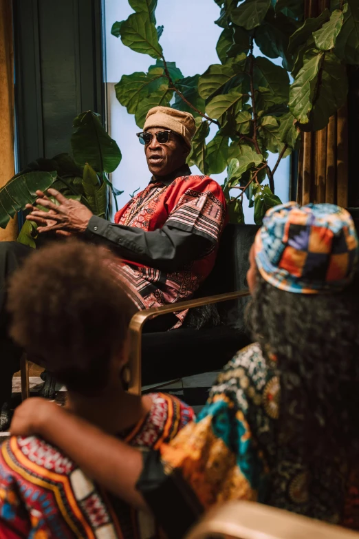 a man sitting in a chair talking to a group of people, a portrait, unsplash, afrofuturism, traditional clothing, miles davis, shot with sony alpha, promotional image