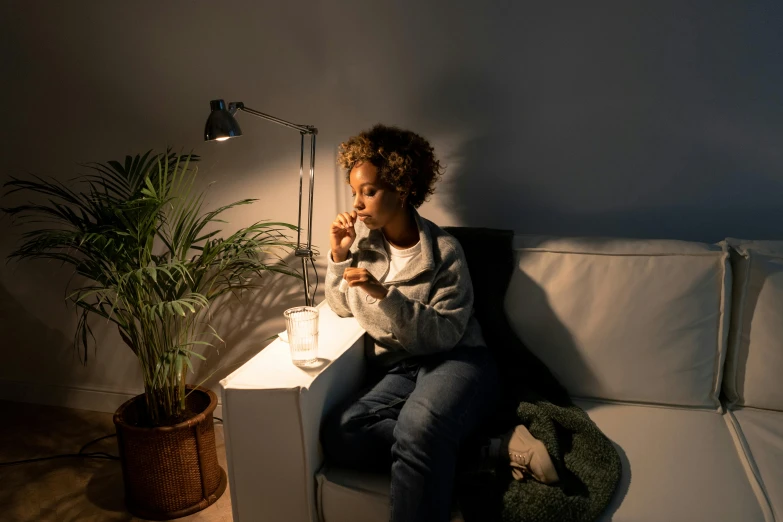 a woman sitting on a couch next to a potted plant, by Niels Lergaard, trending on pexels, night light, girl making a phone call, slight overcast lighting, holding up a night lamp