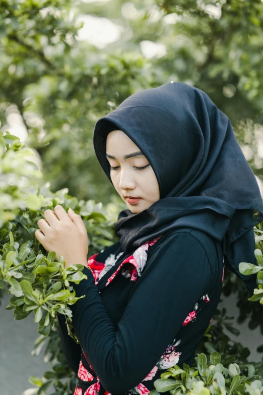 a woman wearing a hijab standing in front of a bush, a picture, inspired by Yu Zhiding, shutterstock, asian human, square, black, cute photo