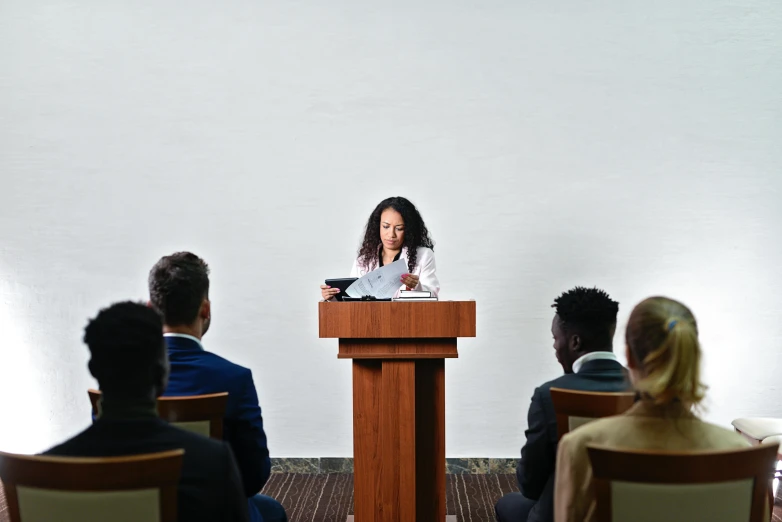 a woman standing at a podium in front of a group of people, pexels contest winner, academic art, plain background, mixed race, sitting at a table, like a catalog photograph