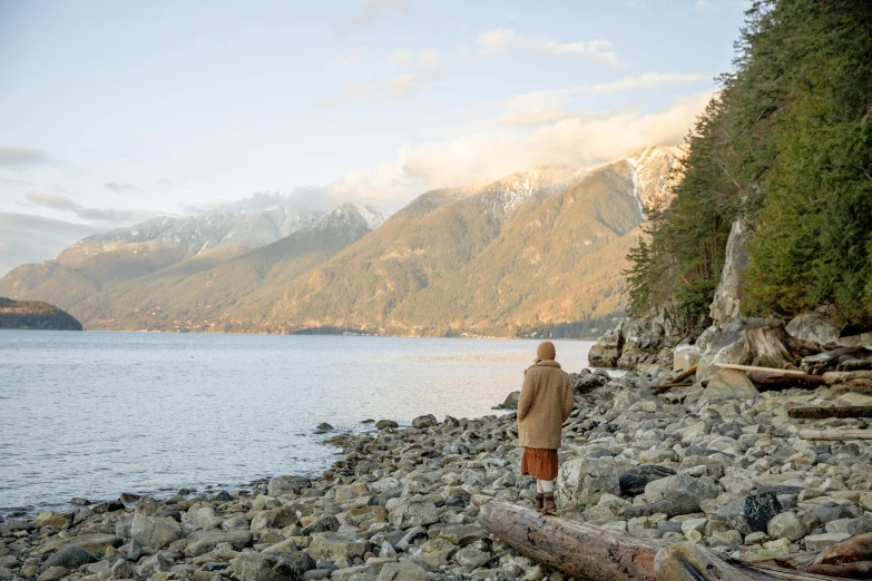 a man standing on a rocky beach next to a body of water, visual art, mountains and oceans, vancouver, brown robes, eleven from stranger things