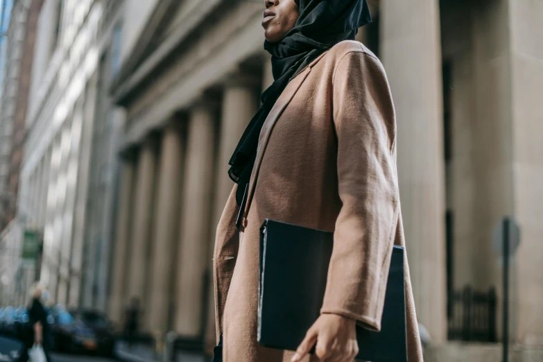 a woman in a hijab is walking down the street, trending on pexels, hurufiyya, holding notebook, wearing a worn out brown suit, black female, thumbnail