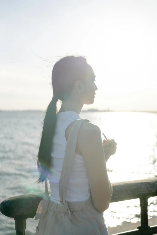 a woman standing on top of a pier next to a body of water, a picture, inspired by Xie Sun, unsplash, long black hair in a ponytail, backlit portrait, overexposed photograph, woman's profile