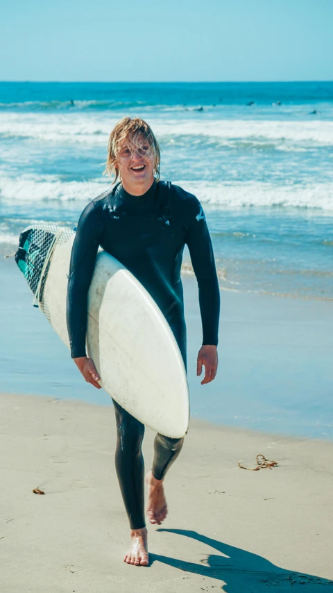 a man walking on a beach holding a surfboard, tony hawk, man in dark blue full body suit, big smirk, corduroy