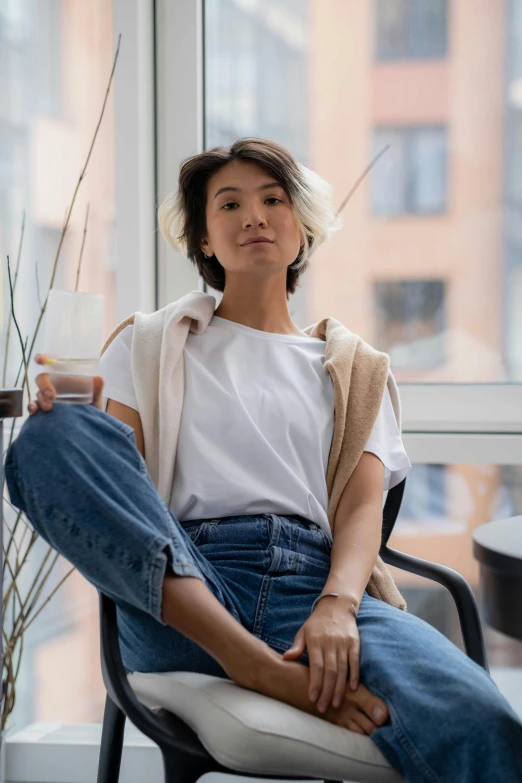 a woman sitting on a chair in front of a window, inspired by Ruth Jên, trending on unsplash, happening, lab coat and tee shirt, asian male, baggy jeans, short platinum hair tomboy