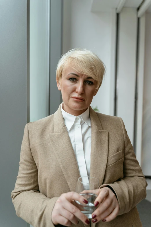 a woman in a suit holding a glass of wine, inspired by Cindy Sherman, bleached blonde short hair, president of belorussia, serious focussed look, faridah malik