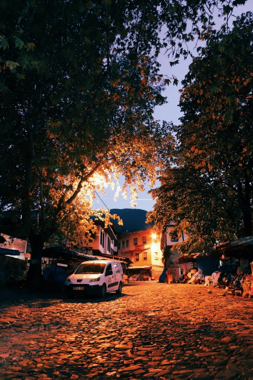 a cobblestone street is lit up at night, by Muggur, happening, sunset with falling leaves, village square, lush surroundings, instagram photo