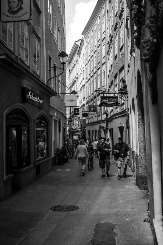 a group of people walking down a street next to tall buildings, a black and white photo, viennese actionism, old shops, 2 2 nd century!!!!! town street, adventuring, shady alleys