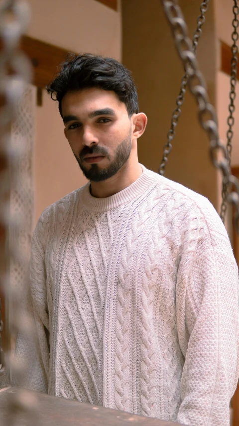 a man standing next to a chain link fence, inspired by Youssef Howayek, pexels, renaissance, wearing a white sweater, vintage clothing, indoor picture, promotional image