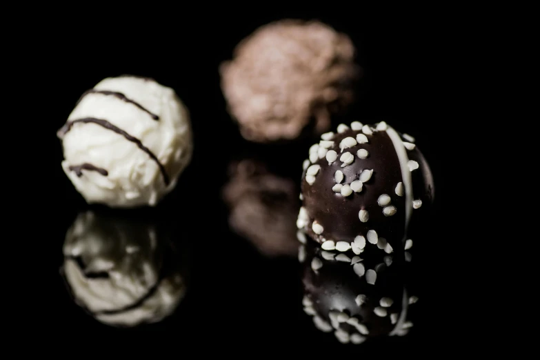 a close up of three chocolate truffles on a black surface, a still life, by Anton Möller, unsplash, hurufiyya, browns and whites, reflections smooth, white and black, ornated