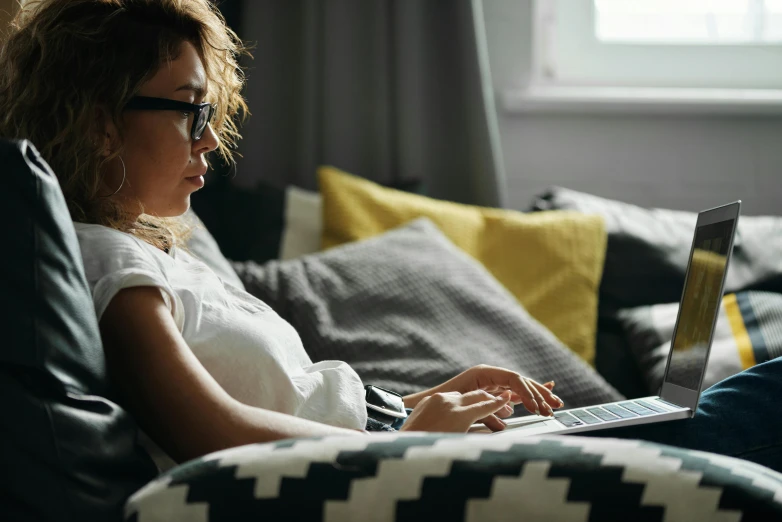 a woman sitting on a couch using a laptop computer, trending on pexels, wearing black rimmed glasses, sitting on edge of bed, cosy, profile image