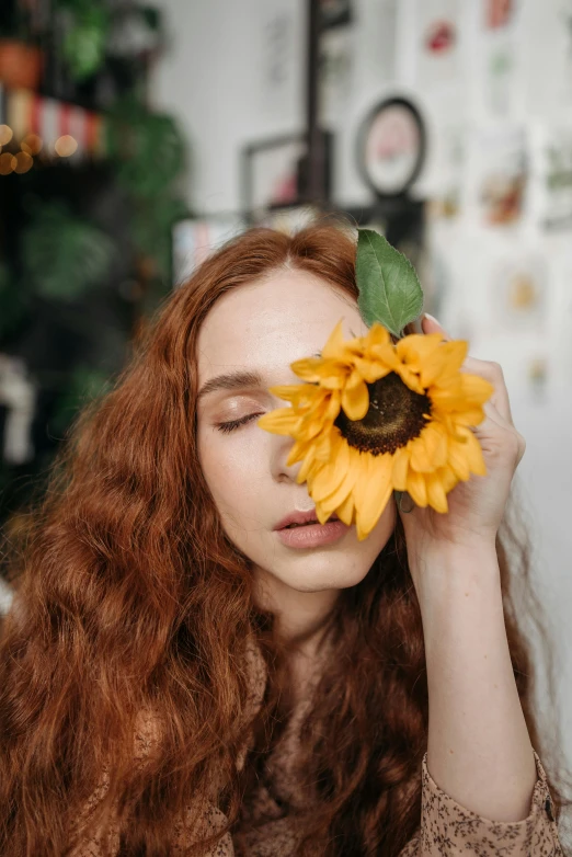 a woman holding a sunflower in front of her face, inspired by Elsa Bleda, trending on pexels, pre-raphaelitism, ginger wavy hair, indoor picture, the girl made out of flowers, eyes half closed