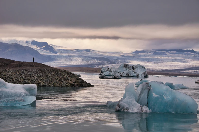 a group of icebergs floating in a body of water, by Þórarinn B. Þorláksson, pexels contest winner, hurufiyya, icy mountains in the background, in muted colours, person in foreground, 2 0 2 2 photo