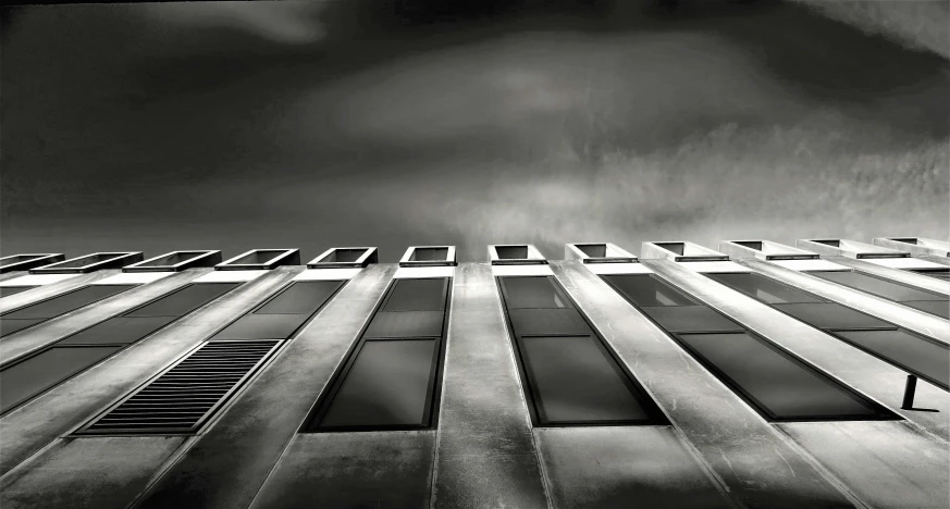 a black and white photo of a tall building, inspired by Pierre Pellegrini, unsplash, brutalism, windows lit up, dramatic grey sky, square lines, silver hues