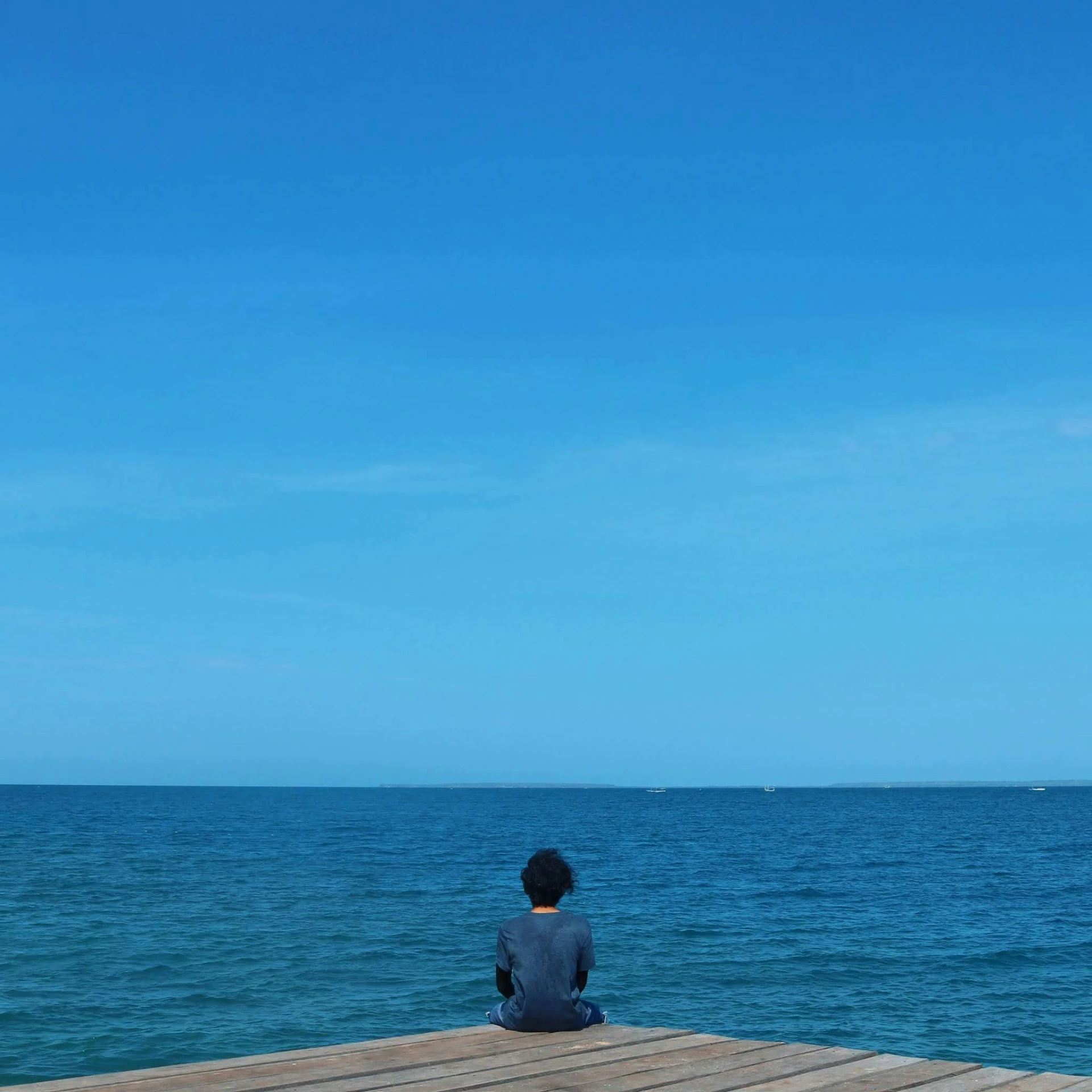 a person sitting on a dock looking out at the ocean, cloudless blue sky, human staring blankly ahead, reddit post, without text