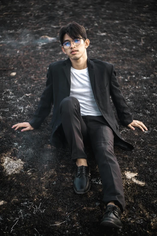 a man in a suit sitting on the ground, an album cover, inspired by Jean Malouel, pexels contest winner, burnt forest, man with glasses, asian male, androgynous person