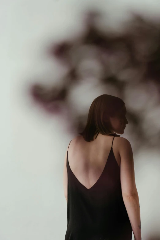 a woman in a black dress standing in front of a tree, inspired by Caro Niederer, trending on pexels, visual art, focus her back, wearing a camisole, dark purple tones, in white room